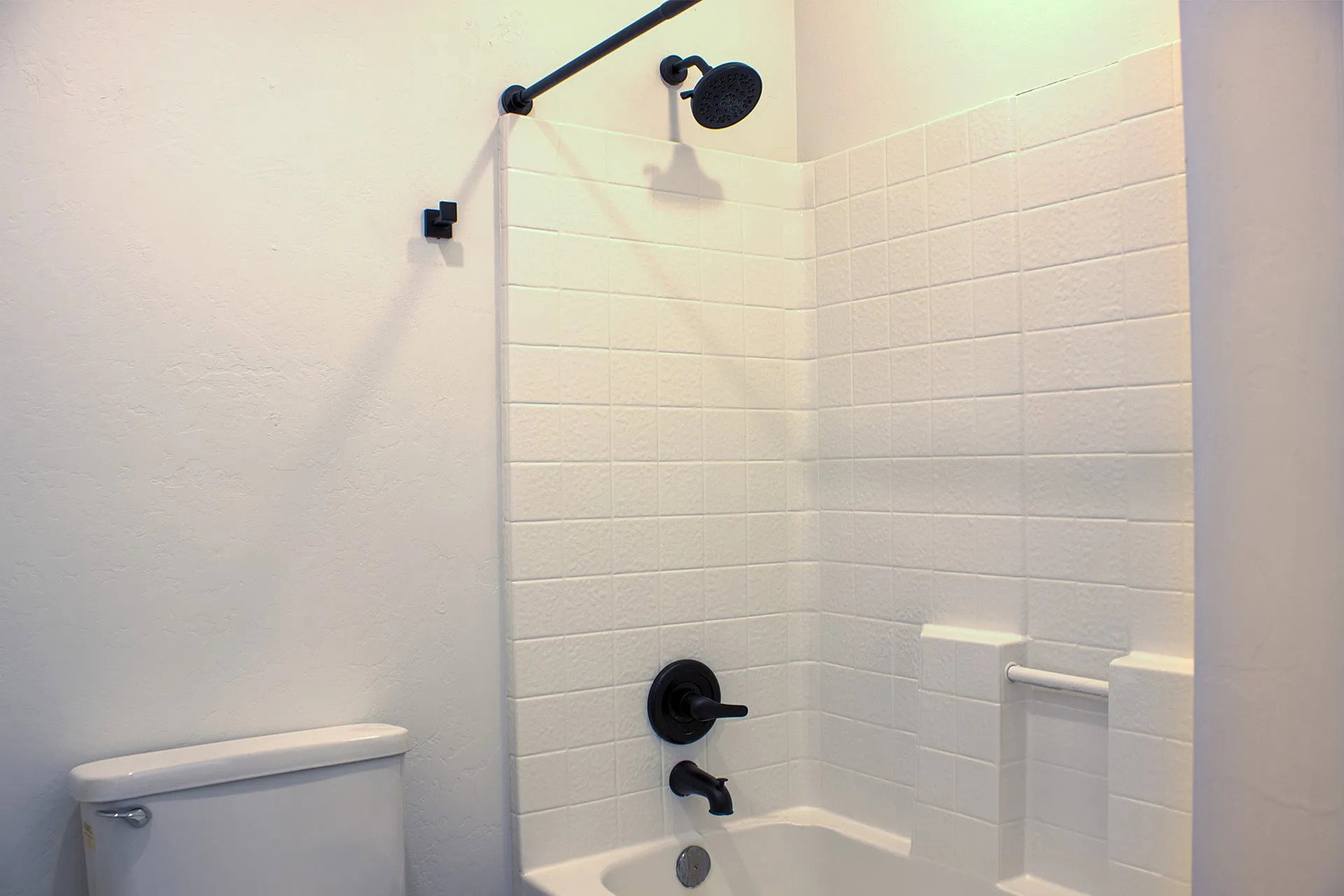 Bathtub and shower combination featuring white tiled walls, matte black shower fixtures, an integrated soap dish, and a white porcelain toilet