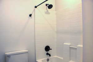 Bathtub and shower combination featuring white tiled walls, matte black shower fixtures, an integrated soap dish, and a white porcelain toilet