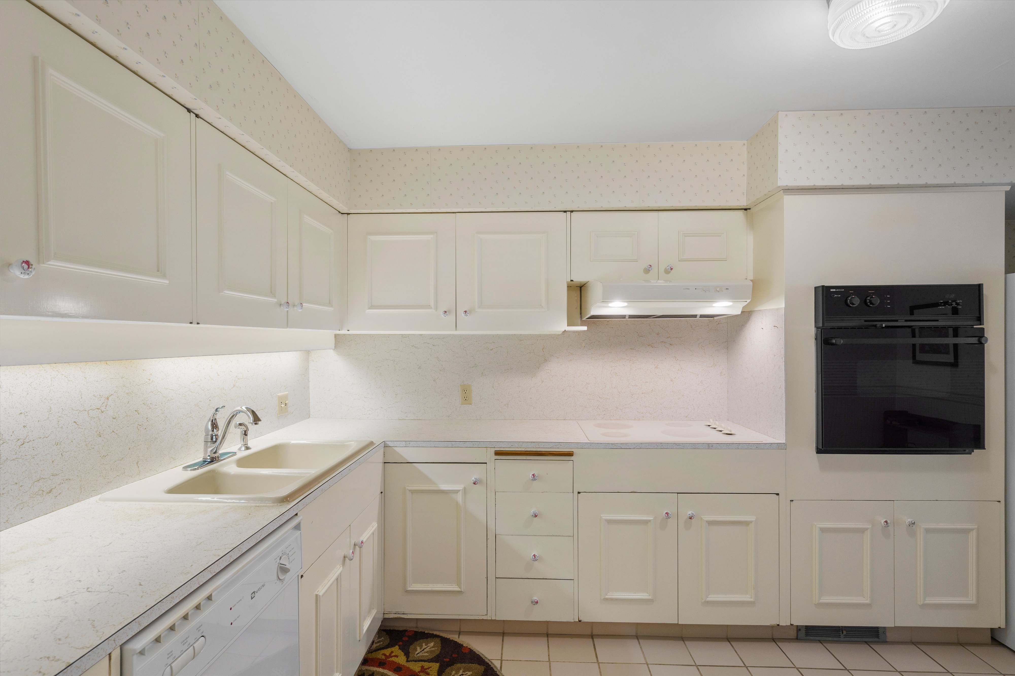 Kitchen featuring white cabinetry with panel detailing, light-toned countertops, a dual basin sink with chrome faucet, a built-in oven, and white tile flooring