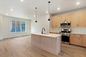 Modern kitchen featuring a central island with a sink, light wood-finish cabinetry, stainless steel appliances, and sleek pendant lighting
