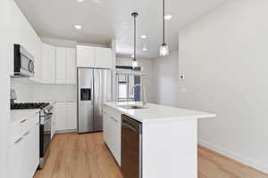 Modern kitchen featuring a central island with integrated sink, stainless steel appliances, white flat-panel cabinetry, a subway tile backsplash, and light wood-finish flooring