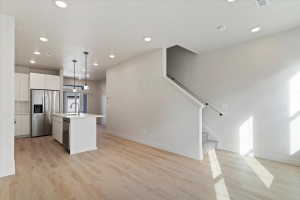 Open concept living area featuring wood-finish flooring throughout, a modern kitchen with white cabinetry and stainless steel appliances, a kitchen island with sink and pendant lighting, recessed ceiling lights, and a carpeted staircase with a white handr
