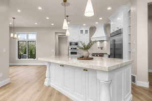 Gourmet kitchen with a large central island featuring a light-toned stone countertop, white cabinetry with brass hardware, a built-in stainless steel refrigerator, and wood-finish flooring