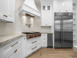 Kitchen featuring white cabinetry, light-toned stone countertops, a stainless steel range with a pot filler, a stainless steel refrigerator, and wood-finish flooring