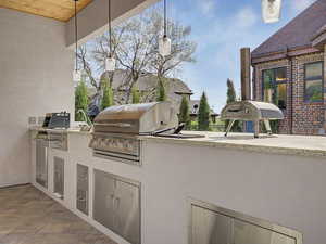 Covered outdoor kitchen featuring a built-in grill, integrated sink, stainless steel cabinetry, stone countertop, and tile flooring
