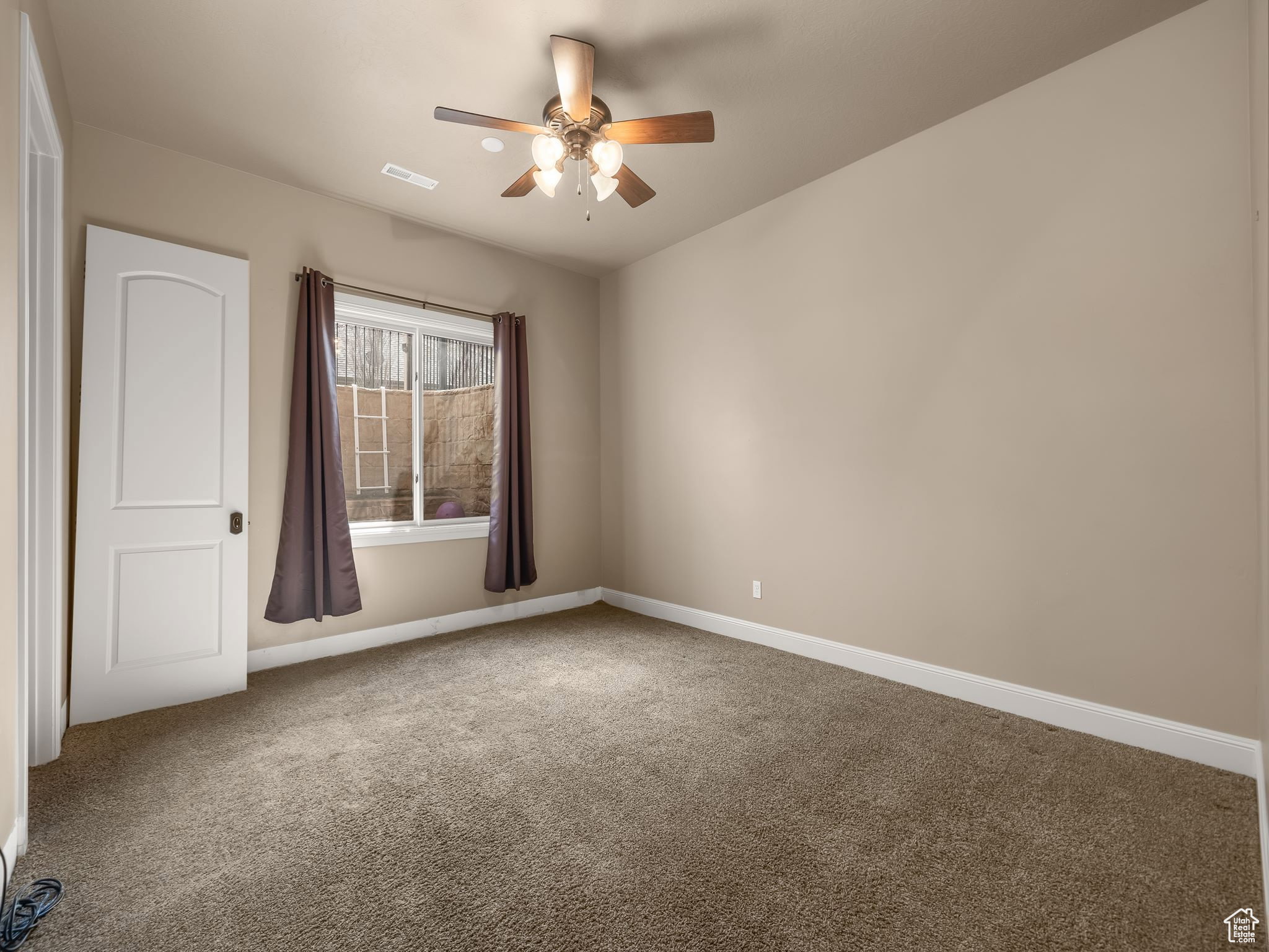 Carpeted room featuring neutral wall tones, a ceiling fan with light fixture, a window with brown drapes, and a white interior door