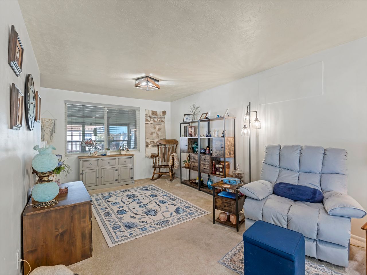 Carpeted living area featuring a large window with horizontal blinds, a ceiling-mounted light fixture, and white painted walls