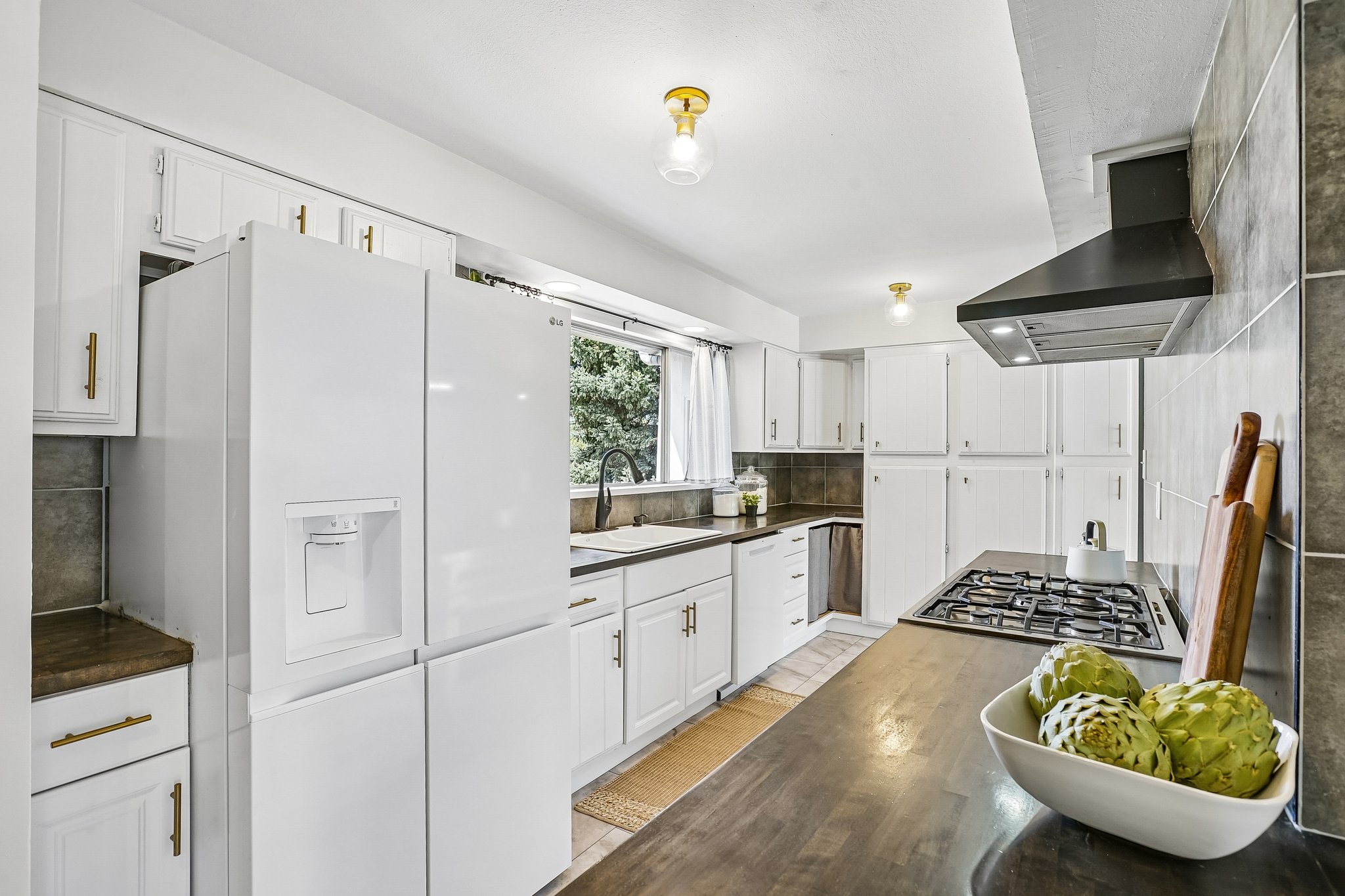 Bright kitchen featuring white cabinetry, brass hardware, dark countertops, a gas cooktop, and a matching black range hood