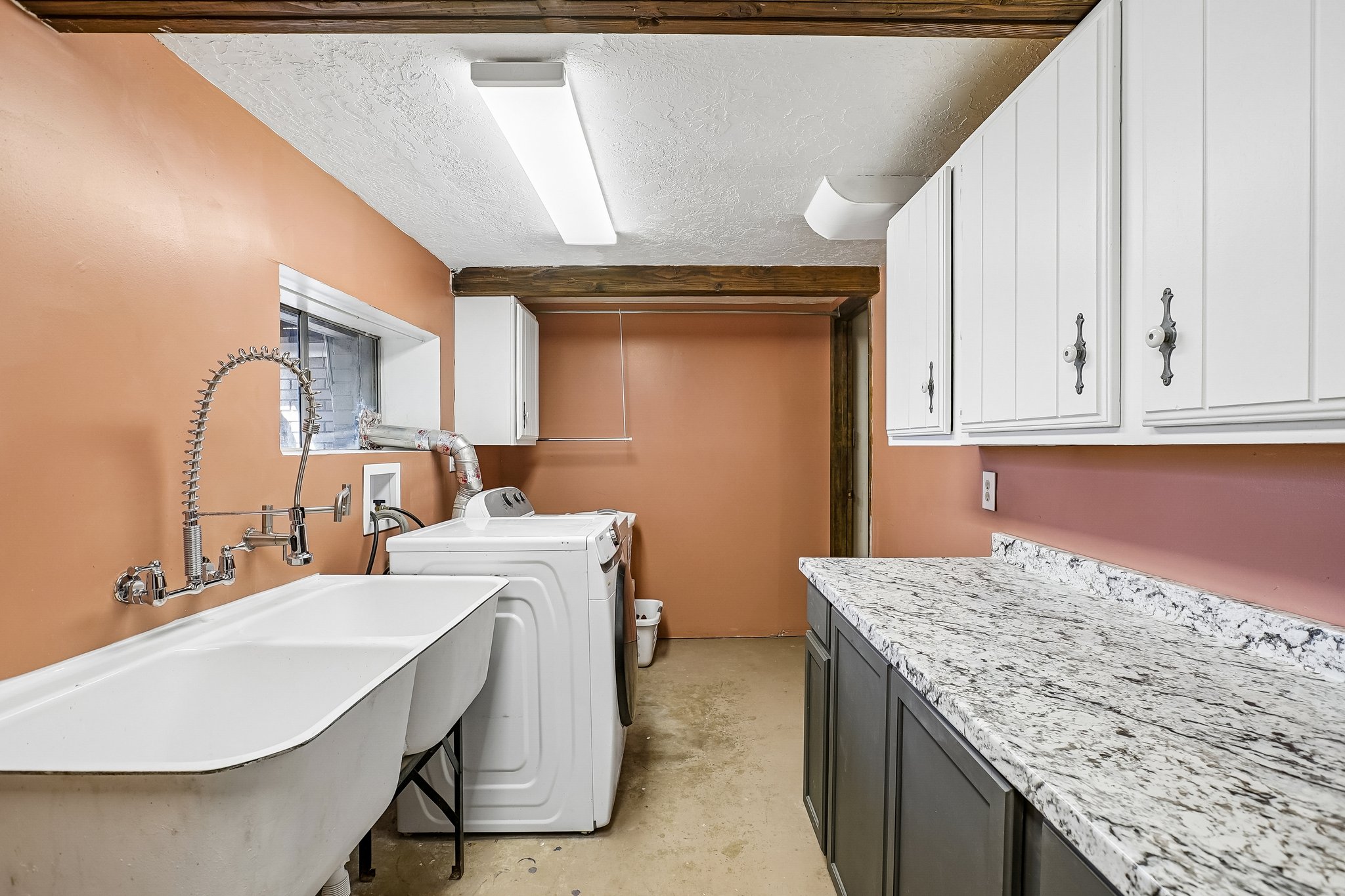 Functional laundry area featuring a utility sink with a spring-spout faucet, a window, exposed ceiling beams, a built-in countertop with dark cabinetry, and upper white cabinetry