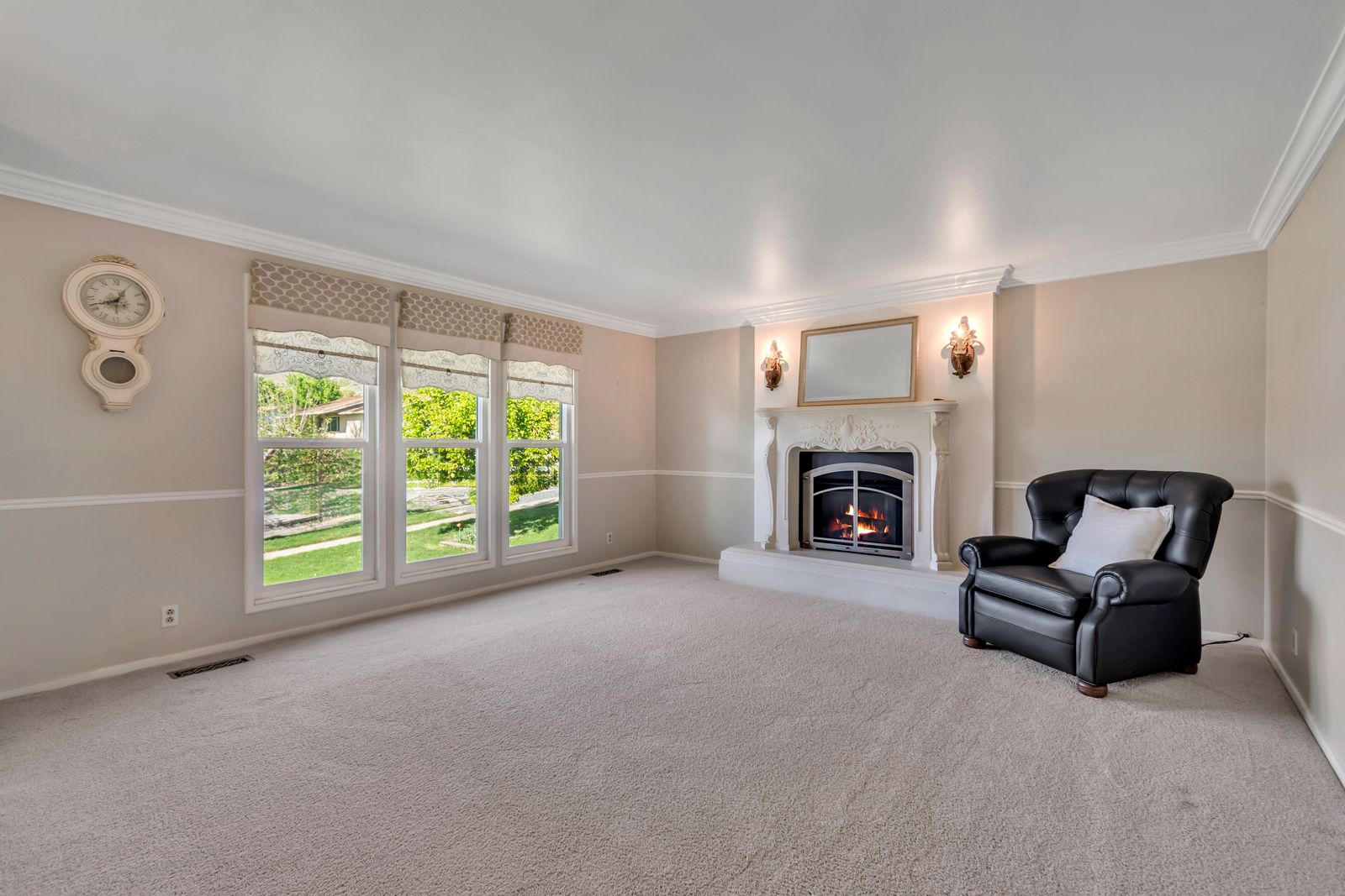 Living area featuring a detailed fireplace with an ornate mantel, crown molding, chair rail molding, three large windows, and wall sconces