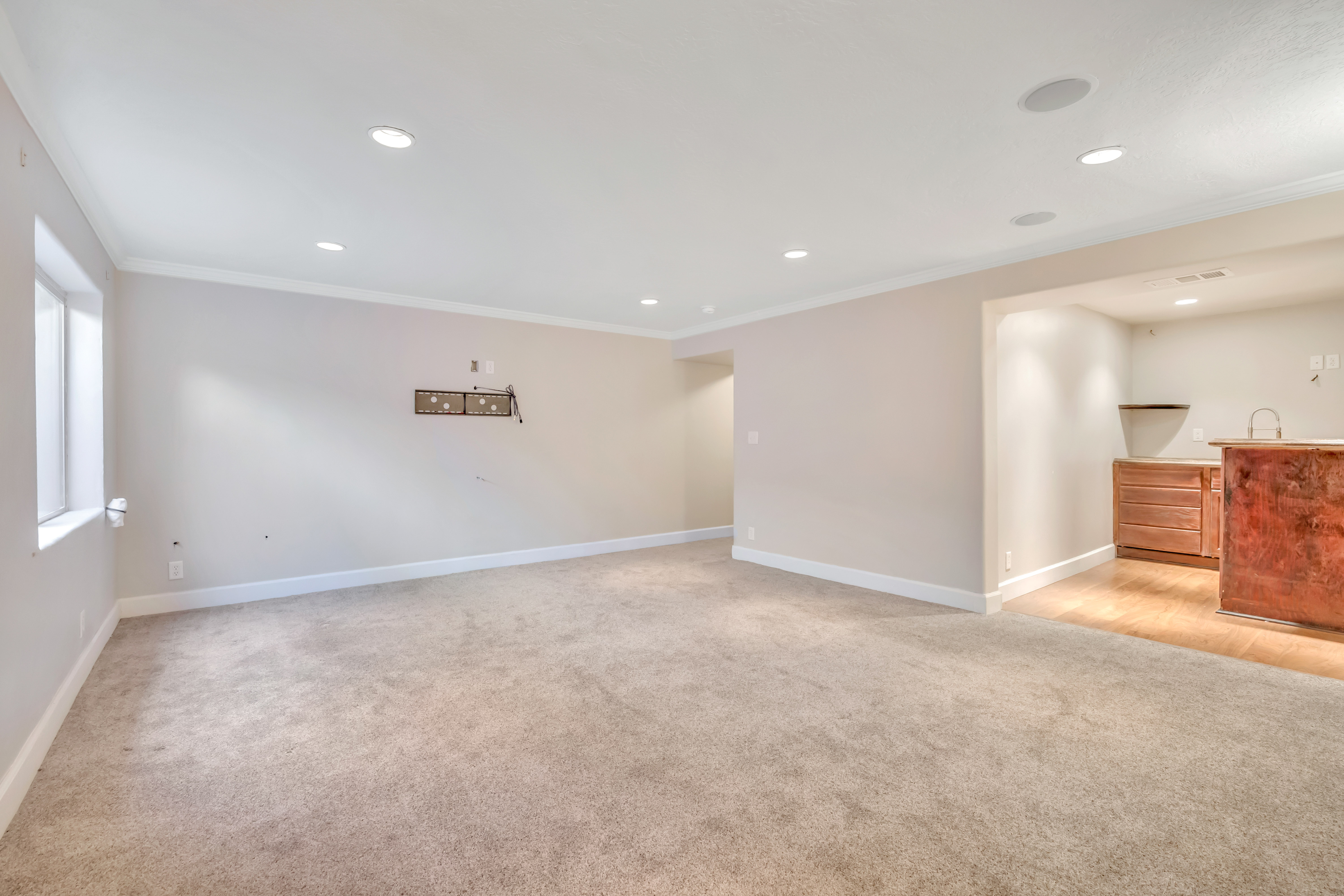 Spacious room featuring neutral carpeting, recessed lighting, crown molding, a window, and a built-in wet bar with wood-finish cabinetry and a sink