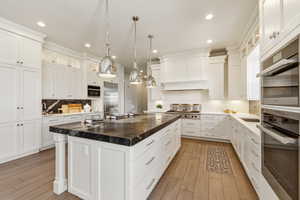 Gourmet kitchen featuring a large central island with a dark stone countertop, white cabinetry with crown molding, stainless steel appliances, and light wood-finish flooring