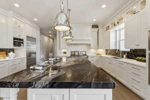 Spacious kitchen featuring a large island with a patterned countertop, white cabinetry, integrated stainless steel refrigerator, built-in microwave, and pendant lighting