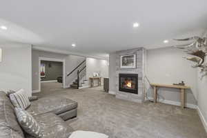 Spacious carpeted room featuring a stone-clad fireplace with a black surround, recessed lighting, white baseboards, and a visible staircase with dark wood handrails and black balusters