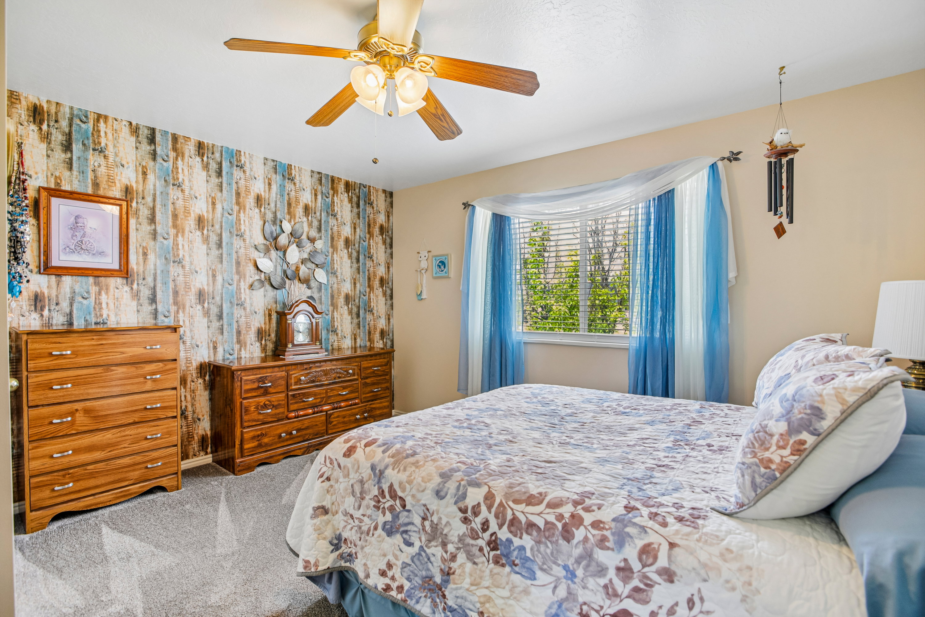Carpeted bedroom featuring a ceiling fan with integrated lighting, a bay window with blinds, and a decorative accent wall