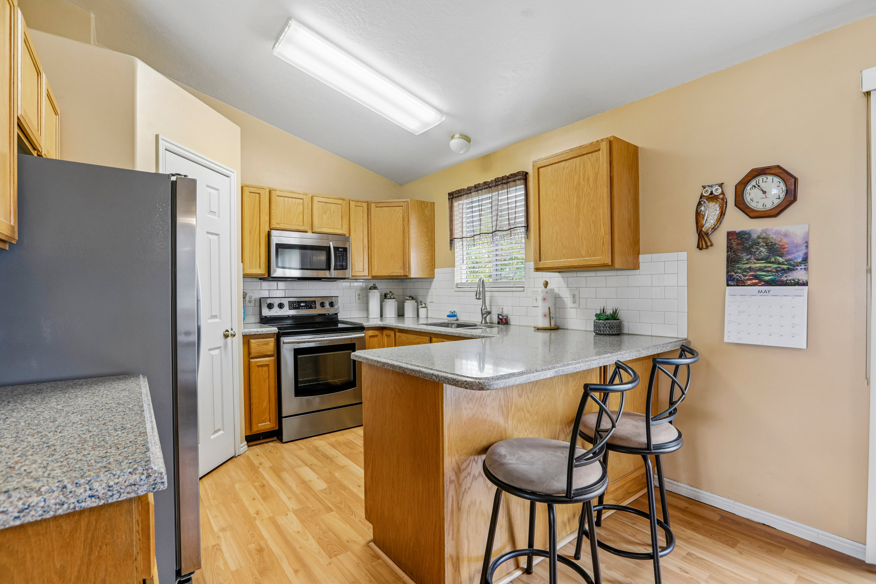 Kitchen featuring wood-finish flooring, light wood cabinetry, stainless steel appliances, a subway tile backsplash, and a peninsula with a stone-finish countertop