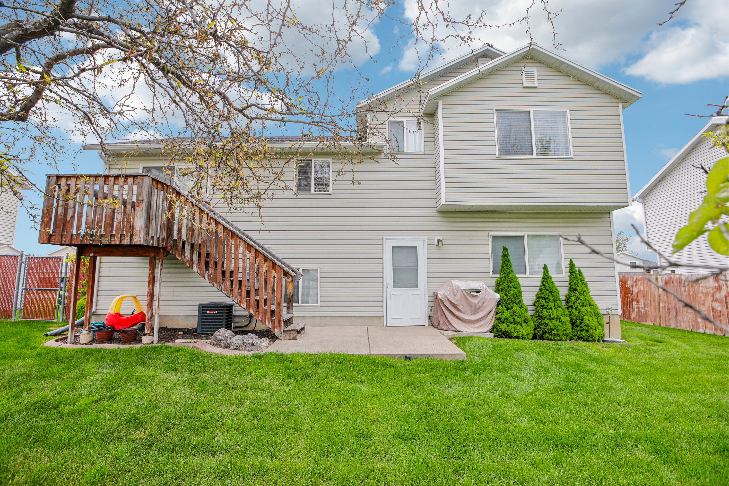 Expansive backyard featuring a manicured lawn, a concrete patio, a wooden deck with stairs, a white exterior door, and three ornamental evergreen shrubs