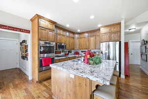 Kitchen featuring a large island with a patterned countertop, wood-finish flooring, stainless steel appliances, abundant wood cabinetry, and recessed lighting