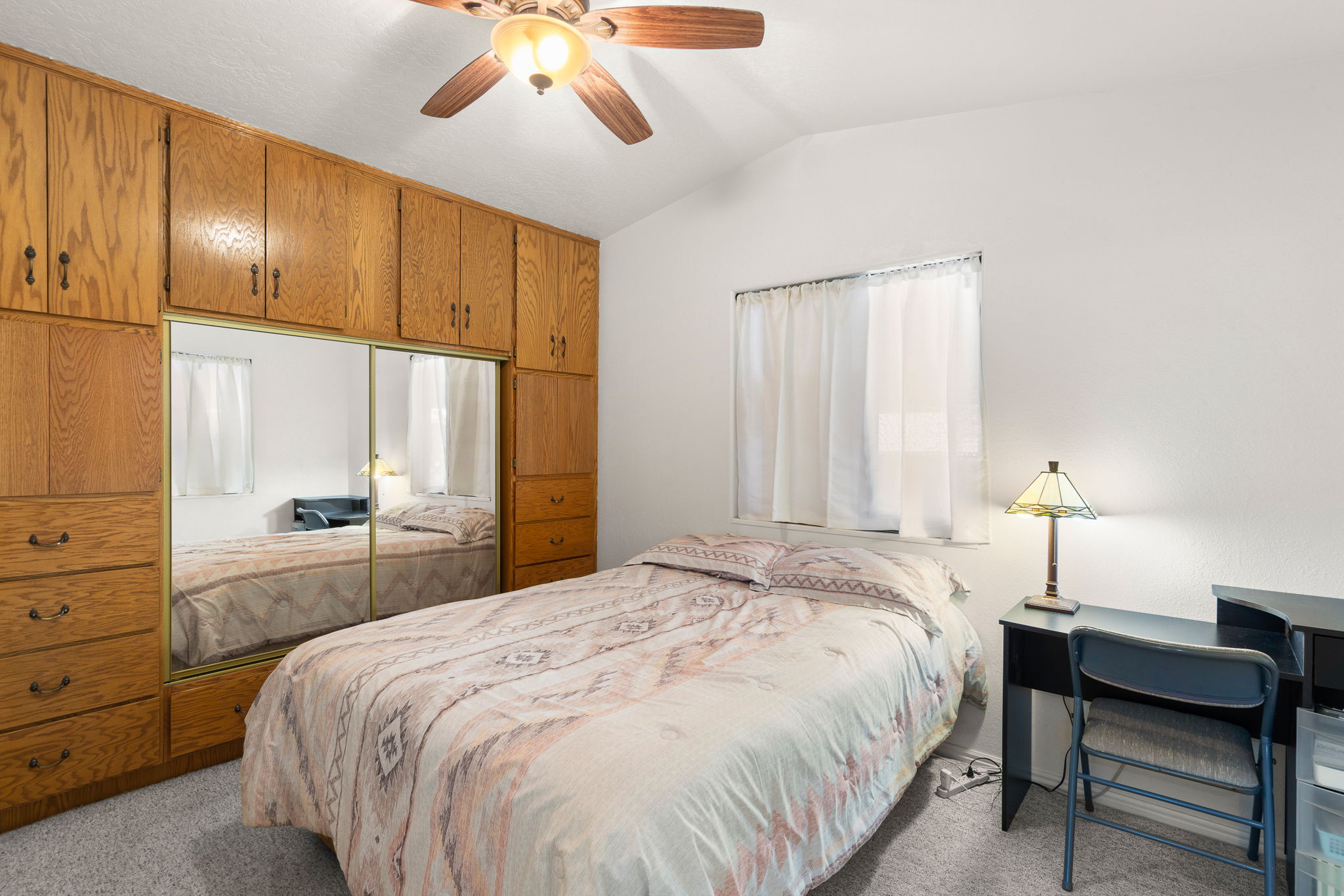 Integrated wood-finish cabinetry with mirrored sliding doors, a white ceiling with a ceiling fan, a window with white curtains, and neutral-toned carpeting