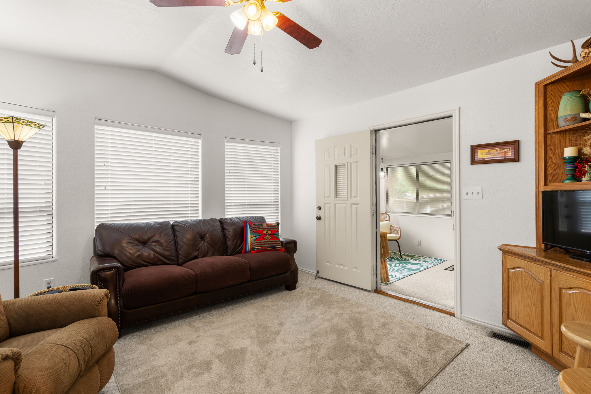 Living area featuring vaulted ceilings, a ceiling fan with light fixture, multiple windows with blinds, plush carpeting, and a built-in wood media console with shelving