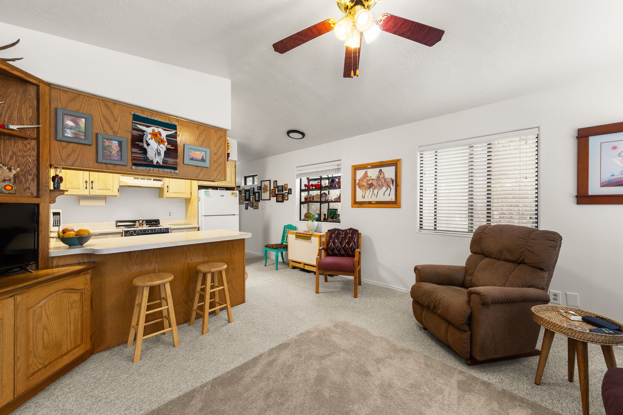 Open-concept living space featuring a kitchen bar, light wood-finish cabinetry, built-in shelving, carpet flooring, and a ceiling fan with integrated lighting