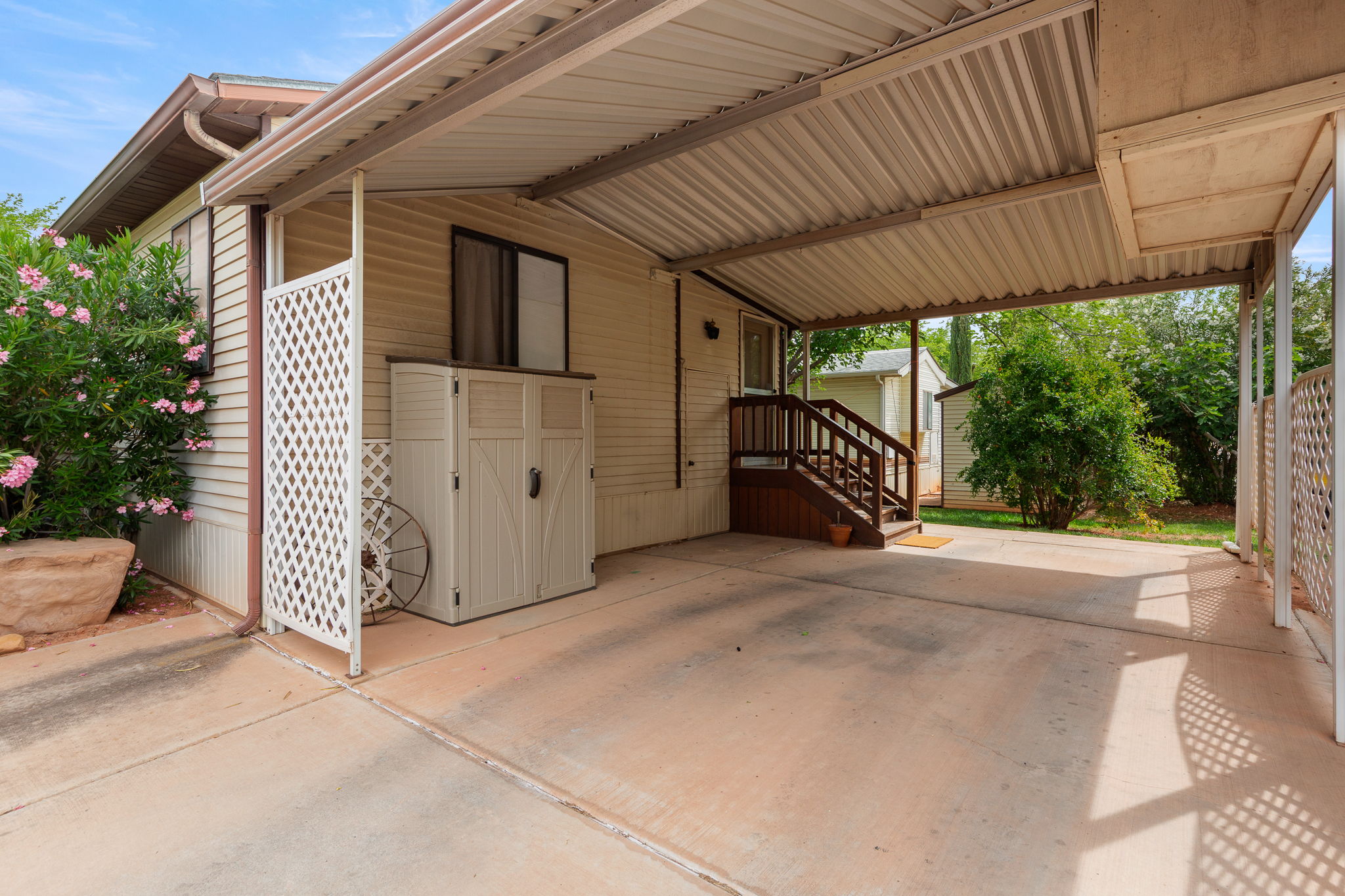 Covered carport with concrete flooring, white lattice privacy screen, and a brown wood-finish porch with stairs