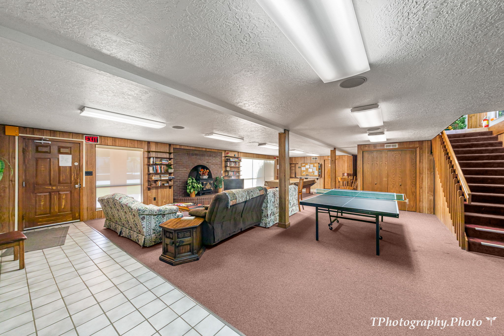 Spacious recreation room featuring wood paneling, a brick fireplace, built-in shelving, carpet flooring, and an interior staircase with wood railings