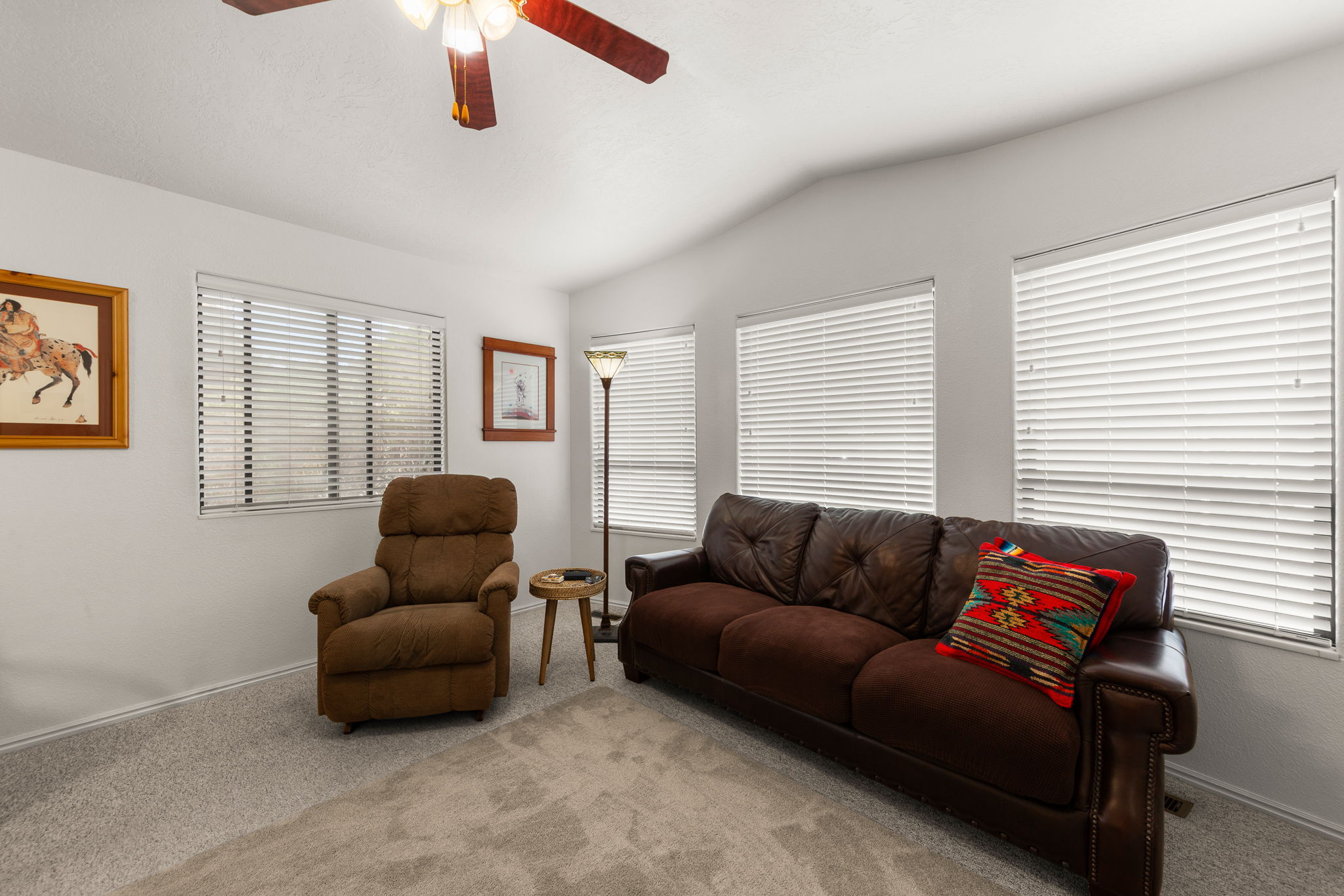 Bright room featuring a vaulted ceiling with a ceiling fan, multiple windows with blinds, and soft gray carpeting