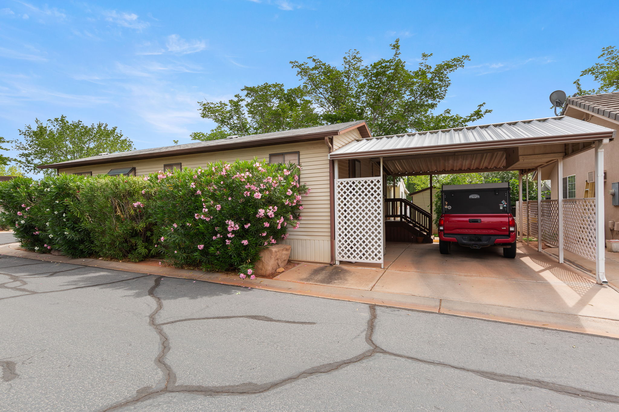 Exterior featuring tan siding, a covered carport, and mature landscaping with flowering shrubs