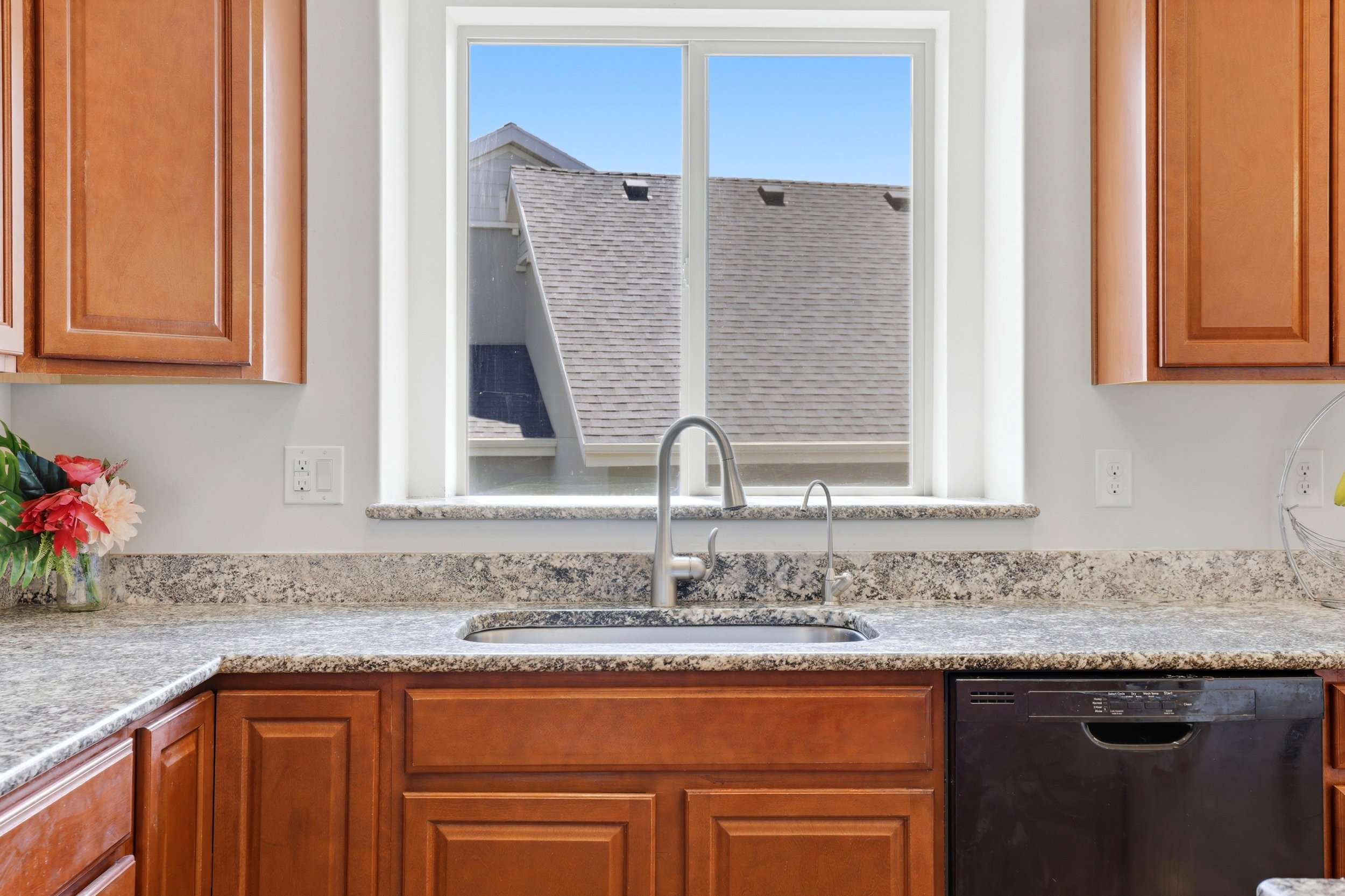 Kitchen workspace featuring a stainless steel sink with a gooseneck faucet, light-toned stone-finish countertops, and wood-finish cabinetry