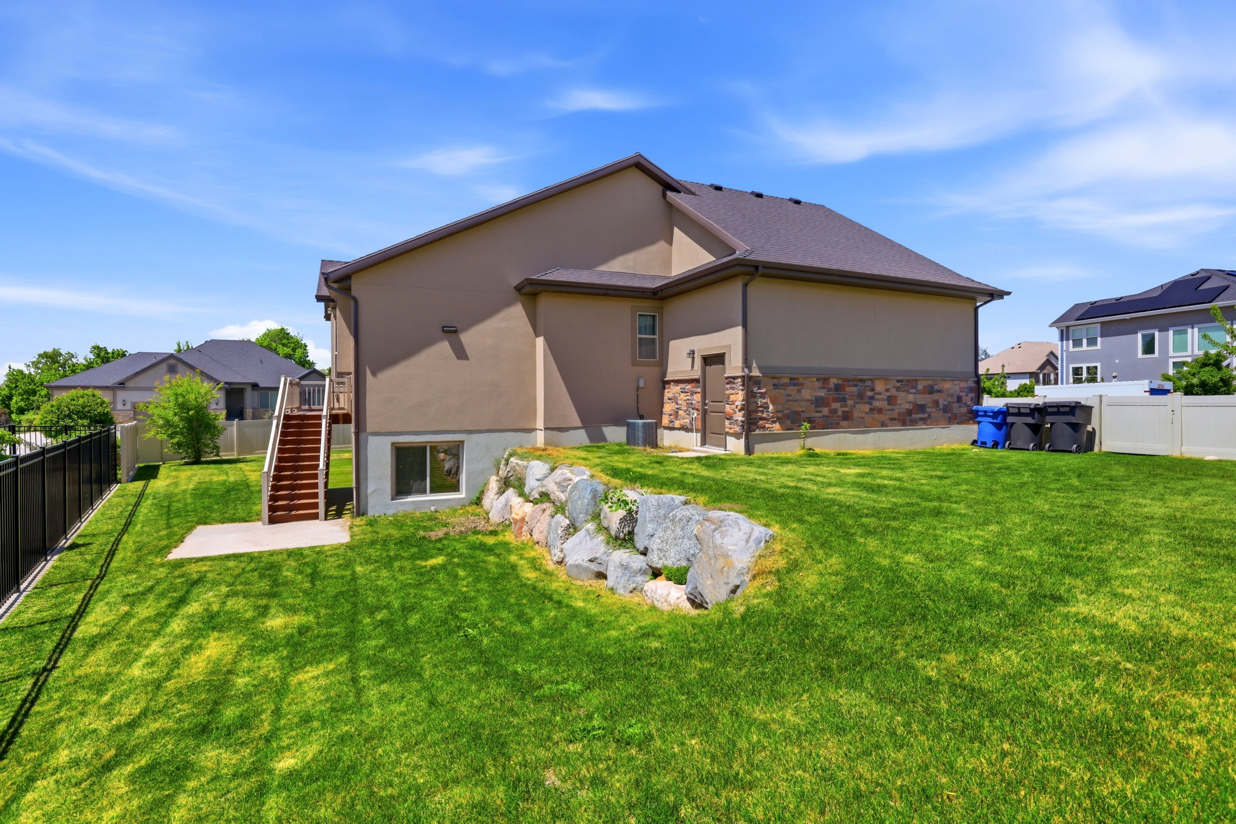 Expansive green lawn surrounds the property, featuring a decorative rock retaining wall, a concrete patio, and exterior stairs leading to a side entry
