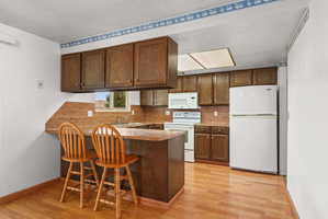 Kitchen featuring wood-finish flooring, dark wood cabinetry, a breakfast bar, white appliances, and an overhead lighting panel