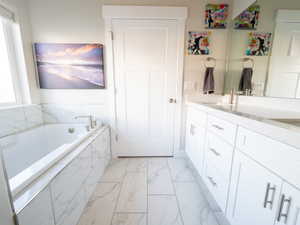 Contemporary bathroom featuring a built-in soaking tub with marble-look tile surround, a spacious double vanity with white cabinetry and light-toned countertops, and marble-look tile flooring