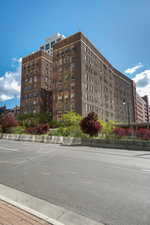 Multi-story brick building with architectural detailing, multiple windows, and a landscaped perimeter featuring mature trees and shrubs