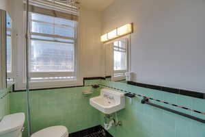 Bathroom featuring mint green wall tiling, black accent trim, a window with horizontal blinds, and a wall-mounted sink with exposed plumbing