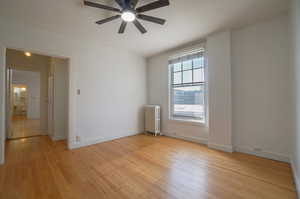 Room featuring wood-finish flooring, a ceiling fan with integrated lighting, a window with horizontal blinds, and a white radiator