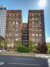 Brick facade building with multiple levels of windows, decorative iron balconies, and mature landscaping at ground level