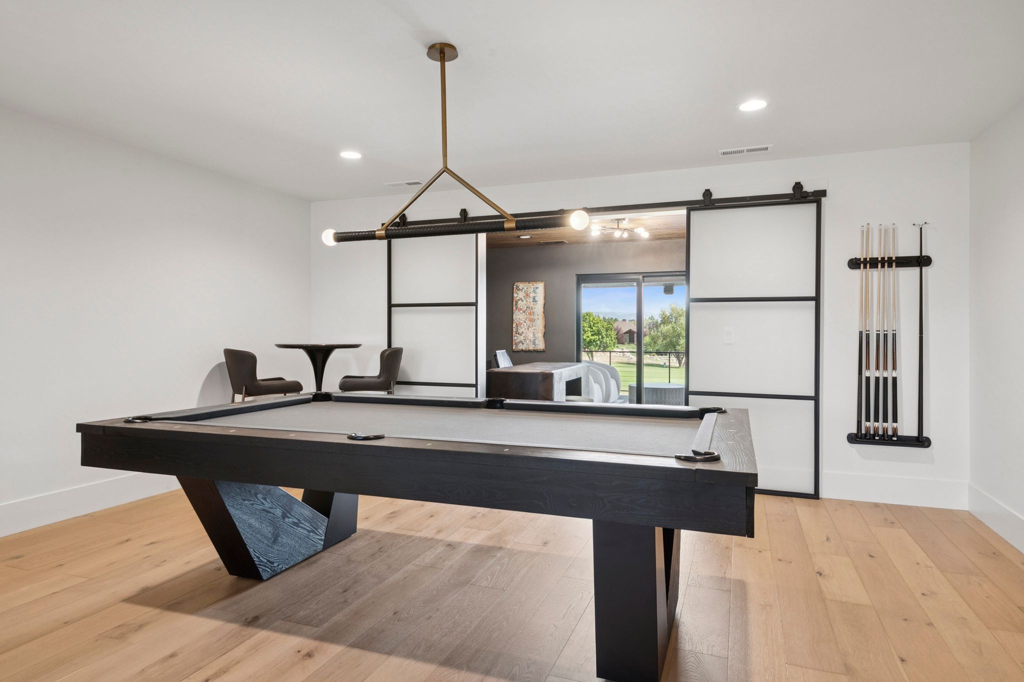 Spacious entertainment area featuring light hardwood flooring, a modern linear chandelier, recessed lighting, a decorative wood slat partition, and black metal stair railing, and frosted sliding doors with access to covered deck.