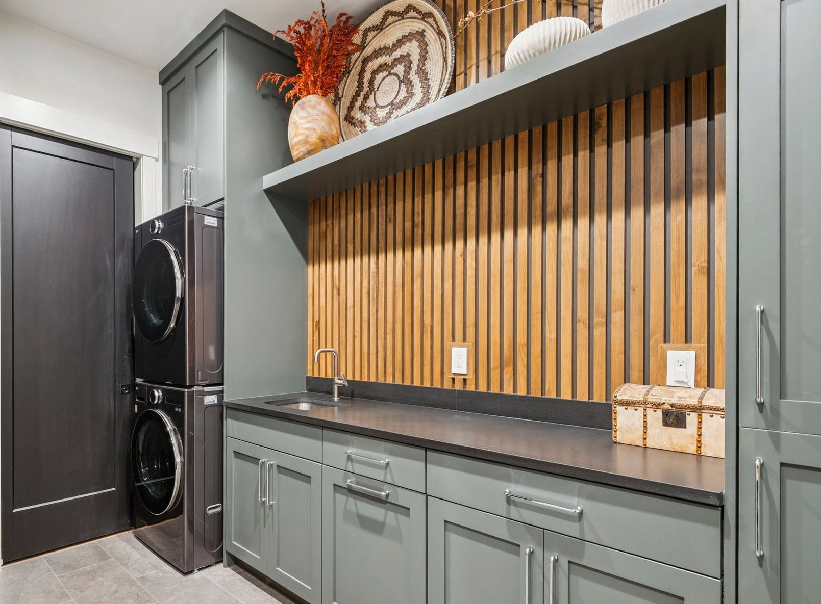 Laundry area featuring stacked LG appliances, custom cabinetry, a utility sink, dark countertop, and a slatted wood accent wall, along with a bench and storage area.
