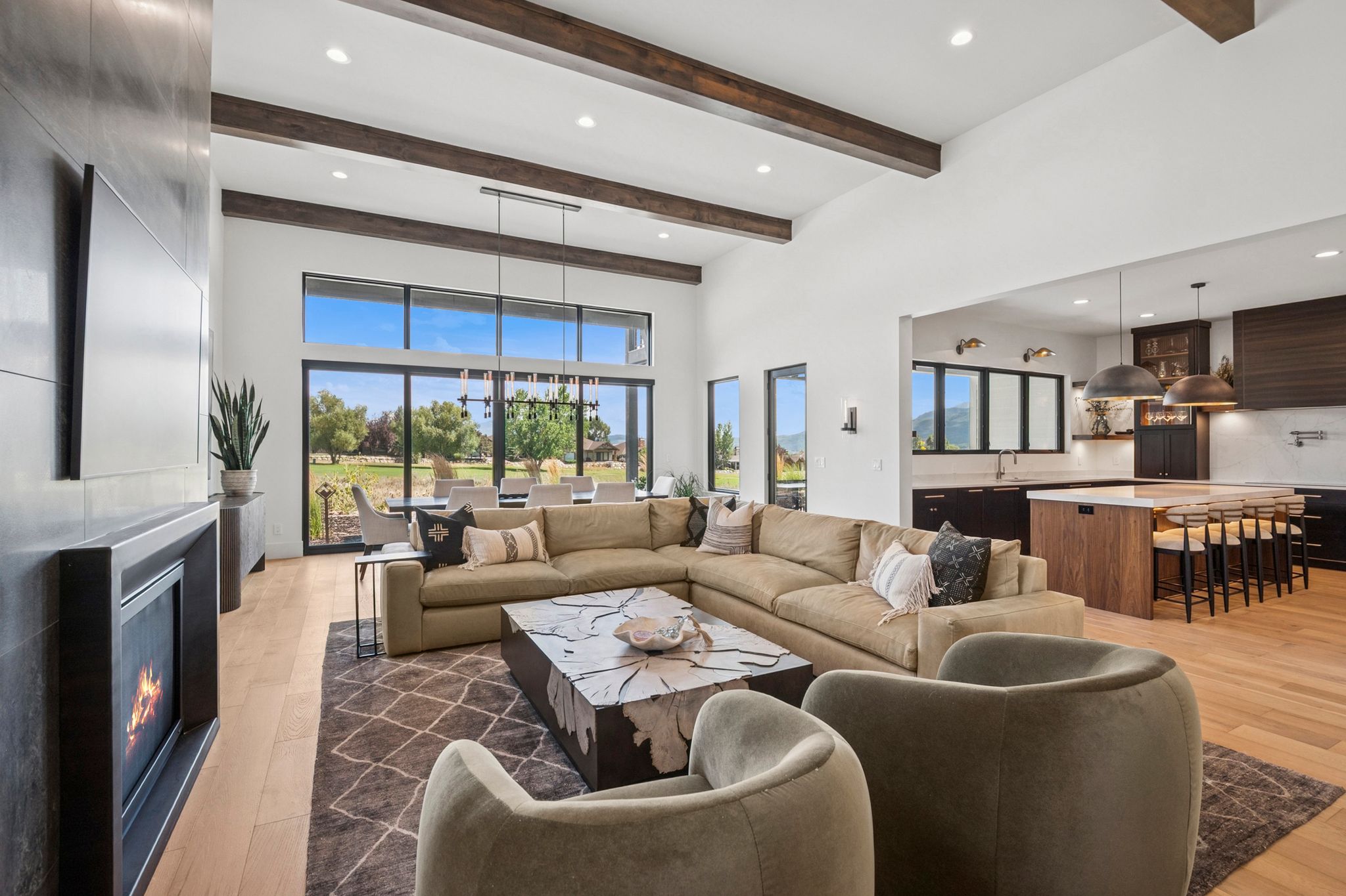 Spacious living area featuring 14' high vaulted ceilings with exposed wood beams, a sleek fireplace with a dark tile surround, and light hardwood flooring.