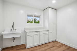 Utility room featuring a wall-mounted laundry sink with a pull-down faucet, white shaker cabinetry, light-toned countertop, and wood-finish flooring