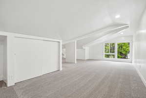 Spacious room featuring light gray carpeting, recessed lighting, white walls, angled ceilings with structural beams, and a window with multiple panes