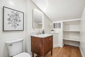 Bathroom featuring a wood-finish vanity with a stone countertop, an illuminated mirror, a white toilet, built-in shelving, and wood-finish flooring