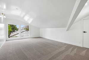 Spacious room featuring vaulted ceilings, a large multi-pane window, flush-mounted ceiling lighting, wall-mounted sconce lighting, and soft grey carpeting