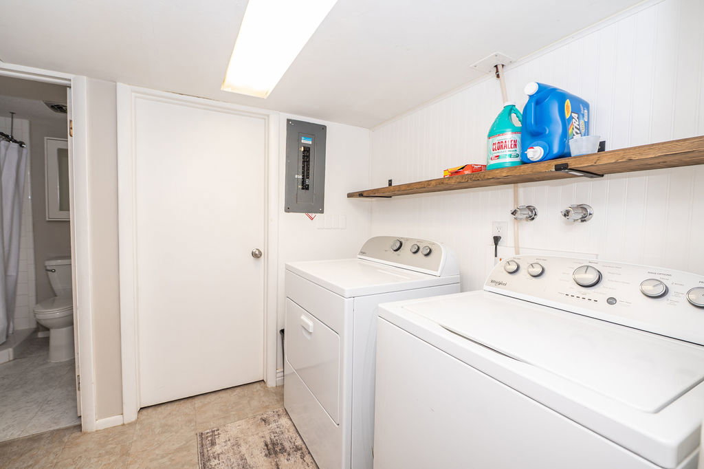 Dedicated laundry area featuring a white top-load washer and dryer, an exposed electrical panel, a wood-finish floating shelf, white plank wall paneling, and overhead fluorescent lighting