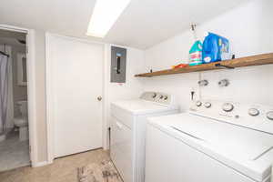 Dedicated laundry area featuring a white top-load washer and dryer, an exposed electrical panel, a wood-finish floating shelf, white plank wall paneling, and overhead fluorescent lighting