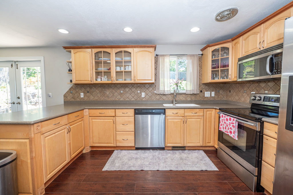 Kitchen featuring light wood cabinetry with upper glass-front cabinets, dark wood-finish flooring, a tiled backsplash, and stainless steel appliances including a dishwasher, range, and microwave
