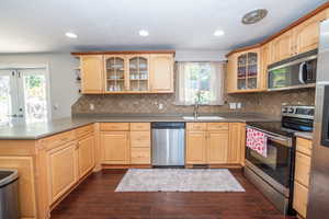 Kitchen featuring light wood cabinetry with upper glass-front cabinets, dark wood-finish flooring, a tiled backsplash, and stainless steel appliances including a dishwasher, range, and microwave