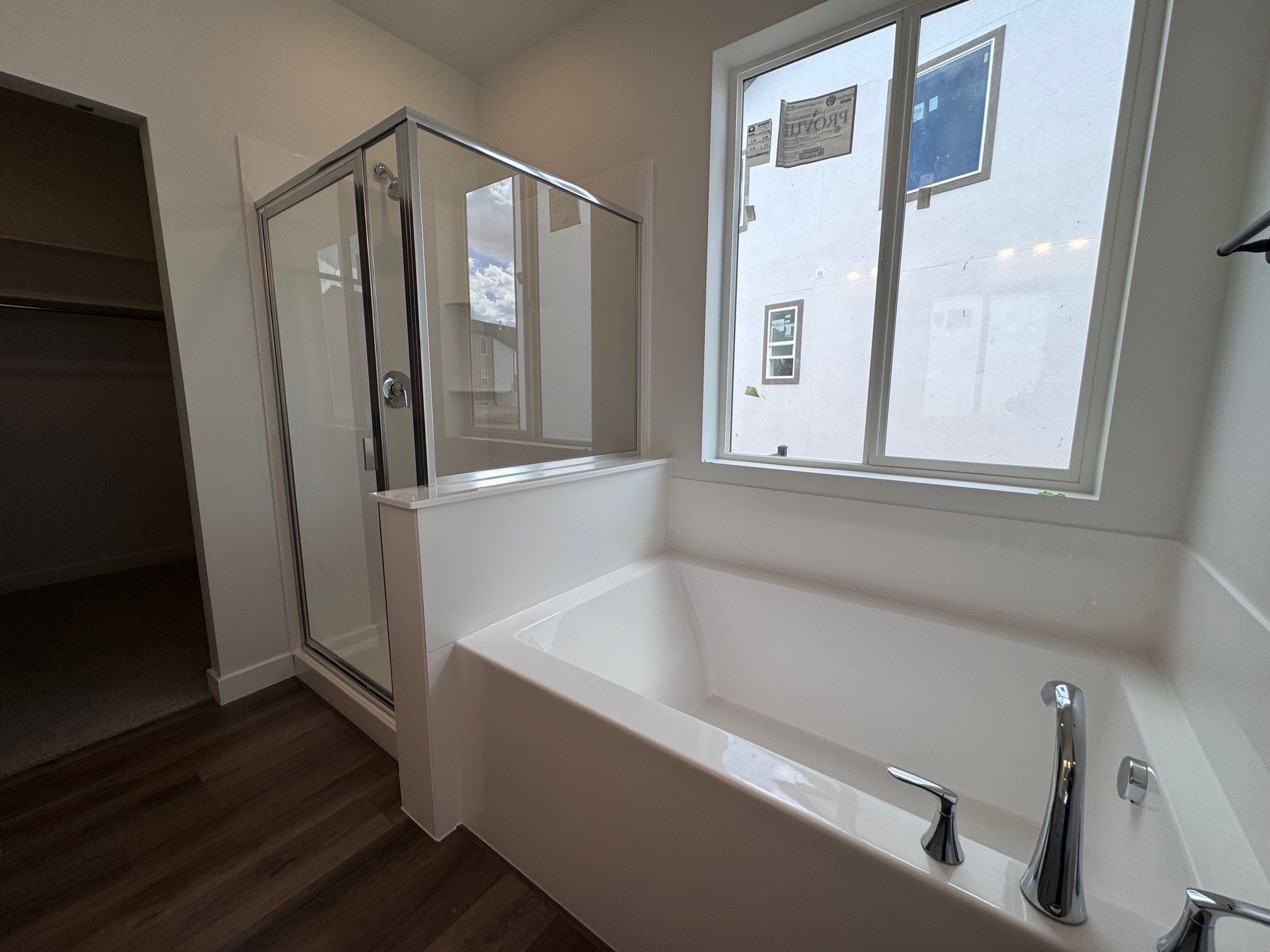 Soaking tub with chrome fixtures, glass-enclosed shower, wood-finish flooring, and a large window