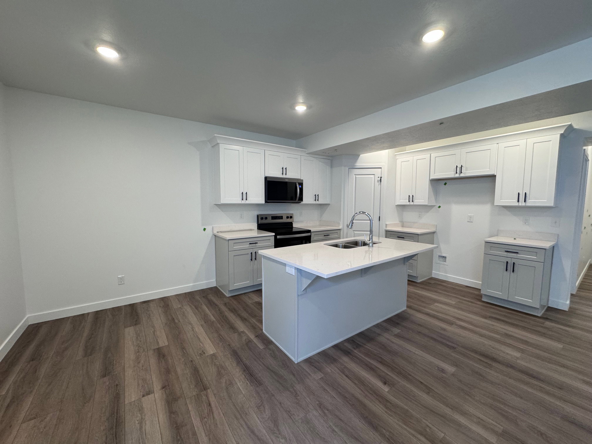 Kitchen featuring wood-finish flooring, recessed lighting, a central island with an integrated sink and gooseneck faucet, light grey base cabinetry, and white upper cabinetry with dark hardware